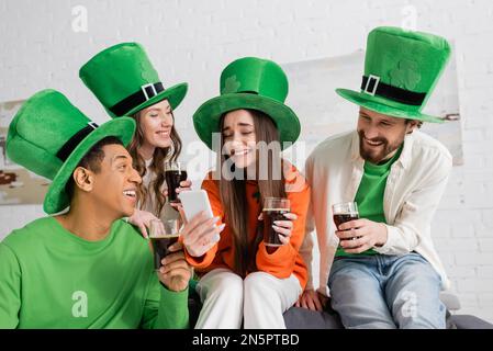 Fröhliche zwischenmenschliche Freunde, die am Saint Patrick Day mit einem Smartphone auf einen Freund schauen, während sie ein Glas dunkles Bier in der Hand halten Stockfoto