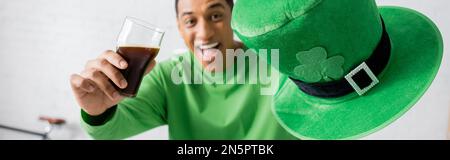 Ein glücklicher afroamerikanischer Mann hält ein Glas dunkles Bier und einen grünen Hut am Saint Patrick Day, Banner Stockfoto