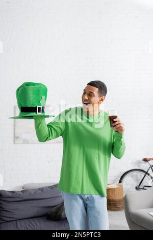 Ein glücklicher afroamerikanischer Mann hielt ein Glas dunkles Bier, während er sich am Saint Patrick Day einen grünen Hut anschaute Stockfoto