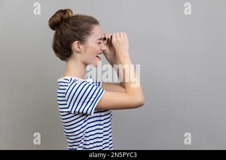 Seitenansicht einer Frau mit gestreiftem T-Shirt, die durch eine monokulare Geste schaut und positive Emotionen ausdrückt, die Sicht zoomt, die Entfernung erkundet. Studioaufnahmen im Innenbereich isoliert auf grauem Hintergrund. Stockfoto