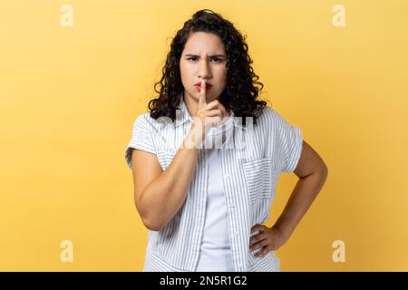 Porträt einer Frau mit dunklem, welligem Haar, die den Finger nahe den Lippen hält, mit einer Psst-Geste, die sie bittet, Ruhe zu bewahren, Privatsphäre zu haben, in die Kamera zu schauen. Innenstudio-Aufnahme isoliert auf gelbem Hintergrund. Stockfoto