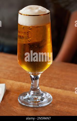 Gekühltes Glas Bier mit Kondensationstropfen auf Glas auf einem Holztisch. Atmosphärisches Bild mit verschwommener Person im Hintergrund. Stockfoto