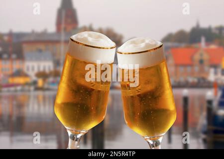 Ein paar Biergläser, die einen Toast ausbringen, während die alte deutsche Stadt im Hintergrund verschwommen ist. Kaltes, erfrischendes Ale mit weißem Schaum. Stockfoto
