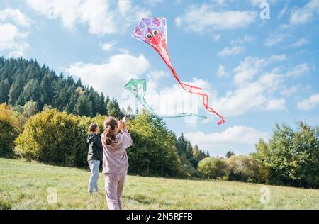 Mädchen und Bruder mit bunten Drachen auf der grünen Wiese in den Bergfeldern. Fröhliche Kindheitserlebnisse oder Zeit im Freien Stockfoto