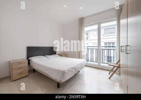 Schlafzimmer mit Eichengarderobe, Balkon mit großen Glas- und weißen Aluminiumtüren, Doppelbett mit schwarzem Holzkopfteil Stockfoto
