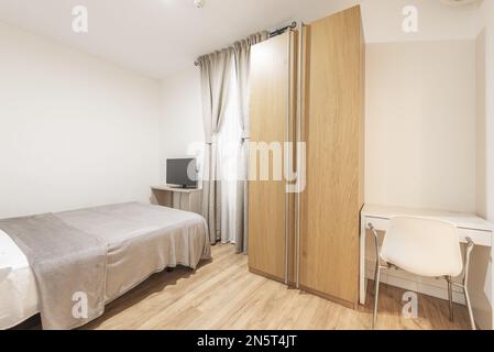 Doppelzimmer mit einem Bett mit einer grauen Bettdecke passend zu den Vorhängen, einem zweitürigen Eichenschrank, einem weißen Schreibtisch mit Stuhl und einem Regal mit Fernseher Stockfoto