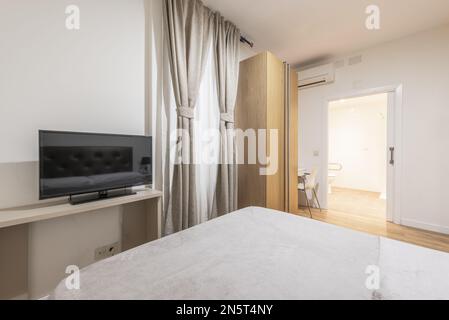 Doppelzimmer mit einem Bett und einer grauen Bettdecke passend zu den Vorhängen, einem Eichenschrank mit zwei Türen, einem weißen Schreibtisch mit Stuhl und einem eigenen Bad Stockfoto