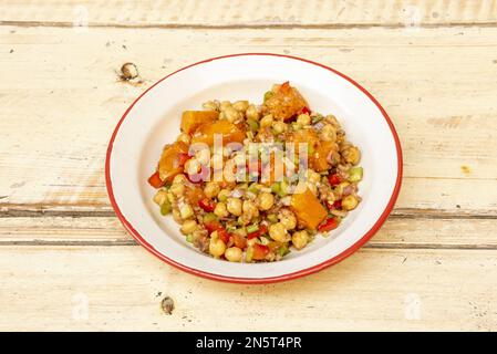 Ein köstlicher veganer Kichererbsensalat mit Kürbis und gemachtem rohem Gemüse in einem rot umrandeten Emaille-Teller Stockfoto