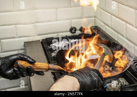 Ein Koch, der zwischen großen, flammenden Rindfleisch-Chop Suey mit einem Wok und einer Edelstahlschöpfchen auf einem Gaskocher sautiert Stockfoto