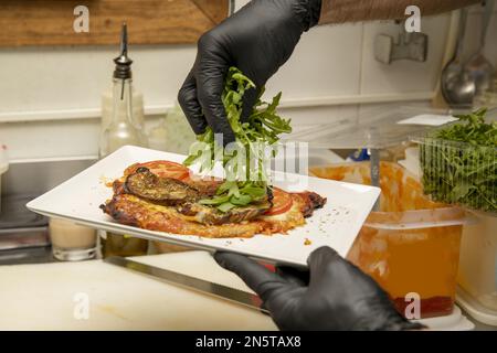 Ein Koch mit schwarzen Plastikhandschuhen, der den letzten Schliff mit Rucola auf einem Schnitzel mit gegrilltem Gemüse in einer Restaurantküche macht Stockfoto