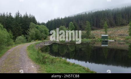 An einem nebligen Tag führt der lange Fußweg Rob Roy Way an einem kleinen See mit einer Fischerhütte und mehreren kleinen Booten vorbei. Die Trossachs, Schottland. Stockfoto