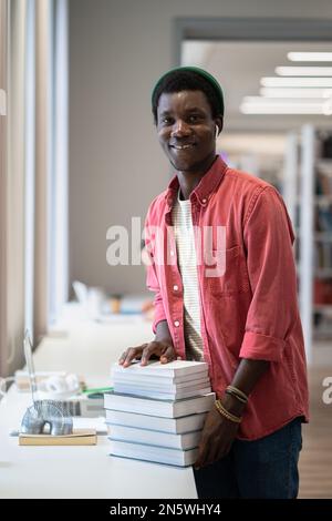 Junger, fröhlicher afroamerikanischer Bibliothekar, der mit einem Stapel Bücher in der öffentlichen Bibliothek steht Stockfoto