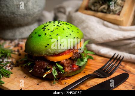 Hausgemachter Burger aus grünem Teig. Grünes Brötchen mit karamellisierten Zwiebeln, einer Scheibe Orangentomate, mit Rucola-Blättern, Barbecue-Sauce, gegrilltem Fleisch. Stockfoto