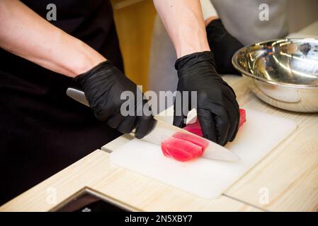 Die Hände des Küchenchefs in schwarzen Handschuhen schneiden die rohen Thunfischfilets mit einem großen, scharfen Messer in Scheiben Stockfoto