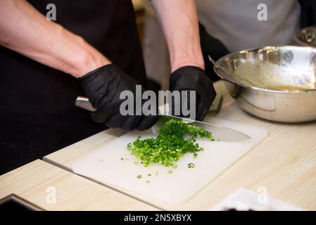Die Hände des Küchenchefs in schwarzen Handschuhen hacken Grüns an Bord, Kochen Stockfoto