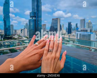 Hände mit Ringen von gerade verlobten und verheirateten Paaren mit einem Bangkok Panorama und Wolkenkratzer vor sich Stockfoto