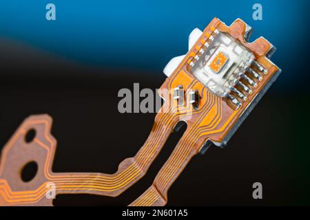 Nahaufnahme des optischen Sensors auf einer elektronischen Leiterplatte oder flexiblen Flachbandkabeln auf dunkelblauem Hintergrund. Kleine orangefarbene Matrize in transparentem Mikrochip. Stockfoto