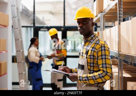 Ein afroamerikanischer Mann, der im Porträt eines Industrielagers auf dem Klemmbrett schreibt. Postlagerpacker trägt einen Helm, verwendet Bestellcheckliste und schaut in die Kamera Stockfoto