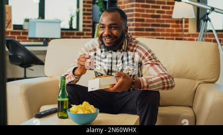 Ein afroamerikanischer Mann, der asiatische Speisen in einer Lieferbox isst, und zu Hause Nudeln mit Stäbchen isst. Ein fröhlicher Kerl, der Spaß hat, eine fernsehsendung zu gucken und Essen zum Mitnehmen auf der Couch zu essen. Stockfoto