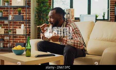 Glücklicher, entspannter Mann, der zu Hause Nudeln isst, vor dem fernseher sitzt und sich einen Film ansieht. Junger Erwachsener, der asiatische Speisen mit Essstäbchen und Flaschen Bier genießt und das Abendessen zum Mitnehmen bestellt. Stockfoto