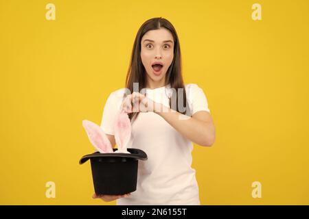 Frohe Ostern. Porträt einer jungen aufgeregten Frau mit Hasenohren isoliert auf gelbem Studiohintergrund. Hasenohren erscheinen aus dem Zauberhut. Stockfoto