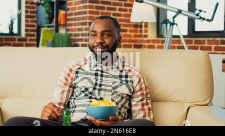 Junge Person trinkt eine Flasche Bier und isst Snacks, und genießt die lieblingssendung im Wohnzimmer. Fröhlicher Kerl, der Spaß mit Film im Fernsehen hat, Burger zum Mitnehmen, Pizza und Nudeln serviert. Stockfoto