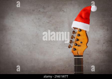 Gitarre mit Weihnachtsmannmütze auf grauem Hintergrund, Platz für Text. Weihnachtsmusik Stockfoto