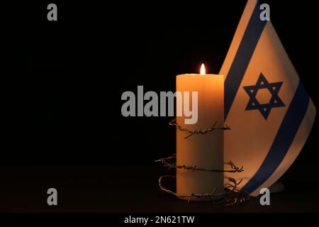 Israelische Flagge, Stacheldraht und brennende Kerze auf schwarzem Hintergrund, Platz für Text. Holocaust-Gedächtnisfeiertag Stockfoto