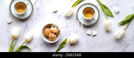 Zarte weiße Tulpen und Teetassen mit herzförmigen Zuckerknochen auf Marmorhintergrund. Draufsicht, Platz für Text kopieren. Stockfoto