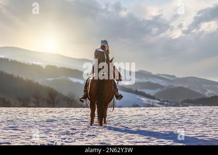 Porträt einer Reitfrau, die bei Sonnenuntergang auf ihrem Pferd vor einer schneebedeckten Winterlandschaft reitet Stockfoto