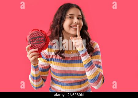Teenager-Mädchen mit Furzkissen, die Stille auf rotem Hintergrund zeigen. Feier zum 1. April Stockfoto