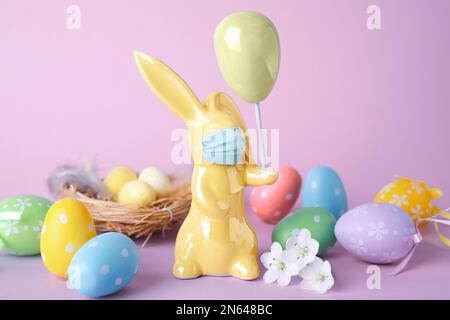 COVID-19-Pandemie. Komposition mit Osterhäschen-Figur in Schutzmaske auf lilafarbenem Hintergrund Stockfoto