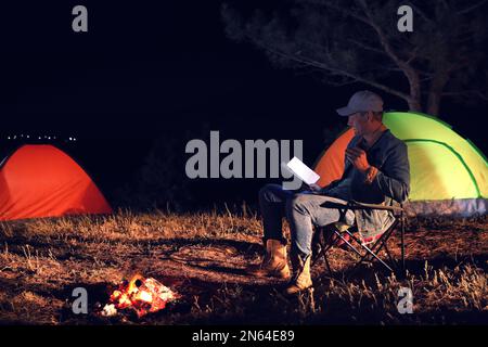 Ein Mann mit Taschenlampe liest nachts ein Buch nahe Lagerfeuer. Campingsaison Stockfoto