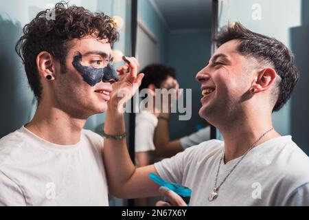 Portrait eines jungen schwulen Paares, das eine Gesichtsmaske aus Ton im Badezimmer aufträgt. Schönheits-Hautpflege-Routine Stockfoto