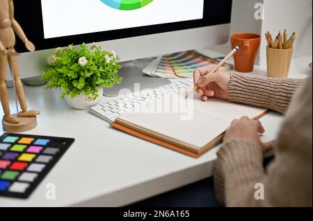 Nahaufnahme einer Grafikdesignerin oder -Redakteurin, die ihre kreativen Ideen auf ihrem Notizbuch aufnimmt und an ihrem Schreibtisch arbeitet. Stockfoto