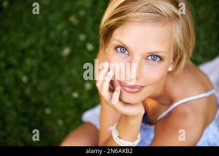 Blauäugige Schönheit. Attraktive junge blonde Frau, die draußen auf dem Gras sitzt und lächelt. Stockfoto