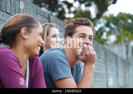 Freundliches Geplänkel zwischen Freunden. Ein Foto von einem Kerl und zwei Mädchen, die gemeinsam lachen, während sie draußen sitzen. Stockfoto