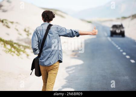 Der abenteuerliche Reisende. Rückansicht eines trendigen jungen Mannes, der am Straßenrand mit einem Auto in der Ferne trampt. Stockfoto