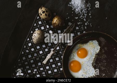 Gebratene Eier in einer Pfanne auf einem Geschirrtuch mit alter Gabel. Frische rohe Wachteleier auf dem Tisch. Draufsicht. Stockfoto