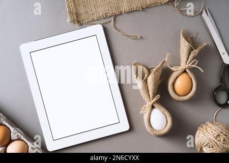 Osterhasen aus Eiern und Servietten auf grauem, hellem Hintergrund. Tischaufnahme mit Tablet-pc mit leerem Bildschirm und osterhaseneiern. Stockfoto