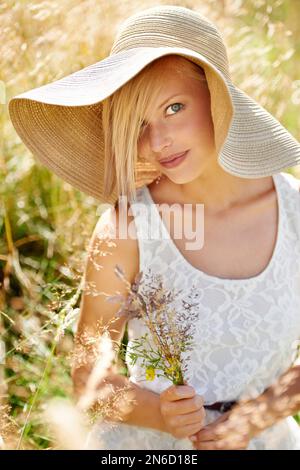 Frische Landschaftspflege. Wunderschöne junge Schönheit, die Wildblumen auf einem Feld pflückt, während sie einen Strohhut trägt. Stockfoto