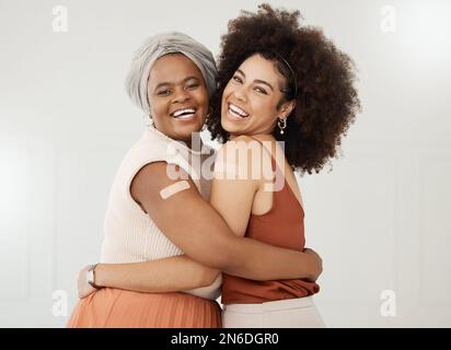 Zwei junge Freunde tragen und zeigen ein Pflaster auf ihren Armen, stehen zusammen auf der Arbeit an einer weißen Wand. Glückliche Geschäftsfrauen, die sich mit A umarmen Stockfoto