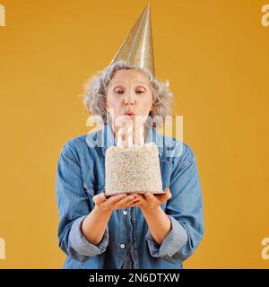 Eine glückliche reife kaukasische Frau bläst Kerzen auf einen Kuchen, den sie hält, während sie einen Geburtstagshut vor einem gelben Hintergrund im Studio trägt Stockfoto