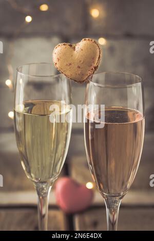 Zwei Gläser mit Sekt und herzförmigen Makronen auf einem rustikalen Holztisch, romantische Atmosphäre, vertikal Stockfoto