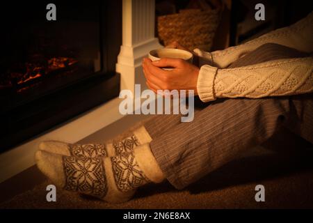 Eine Frau in warmen Socken, die ein heißes Getränk am Kamin hält, mit brennendem Holz zu Hause, Schließung Stockfoto