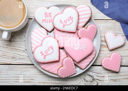 Dekorierte herzförmige Plätzchen und Kaffee auf weißem Holztisch, flach liegend. Valentinstag-Leckerbissen Stockfoto