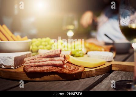 Nahaufnahme einer Vielzahl von Snacks auf einem Tapas-Holzbrett draußen auf einem Tisch. Käse, Brot, frische Trauben und Aufschnitt zum Mittagessen auf einem Weinberg Stockfoto