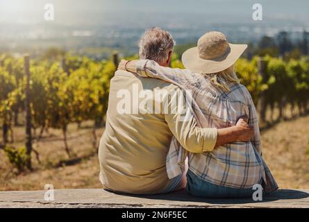 Rückblick auf ein unbekanntes Seniorenpaar, das auf seiner Farm mit den Armen umeinander sitzt. Kaukasische Bauern, die sich anfreunden, während sie ihren Weinberg bewundern und Stockfoto