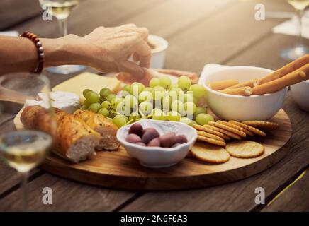 Eine unbekannte Frau, die eine Vielzahl von Snacks auf einem Tapas-Holzbrett draußen nahm. Käse, Brot, frische Weintrauben und Aufschnitt zum Mittagessen arrangiert Stockfoto