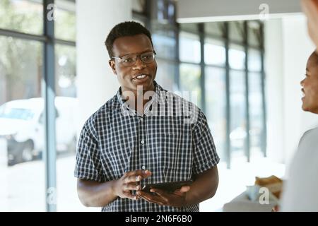 Junger, glücklicher afroamerikanischer Geschäftsmann, der ein digitales Tablet in der Hand hält, während er mit Kollegen in einem Büro spricht. Männlicher Boss mit Brille und Stockfoto